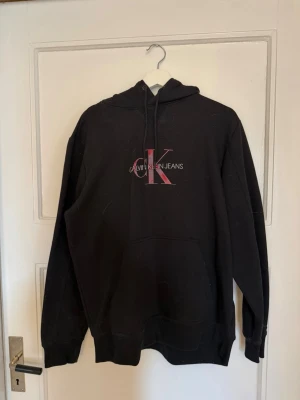 Svart hoodie från Calvin Klein Jeans - Svart hoodie från Calvin Klein Jeans med stor CK-logga och text på bröstet i vitt och rött. Klassisk modell med huva och dragsnören samt ribbade muddar vid ärmar och nederkant. Perfekt för en avslappnad och stilren look. Lite slitet märke 