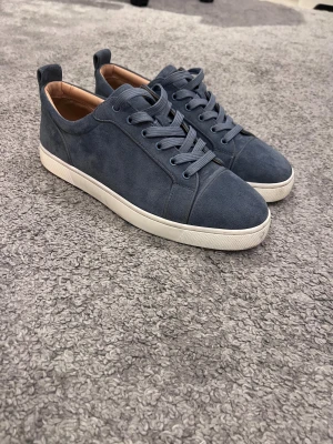 Grå mocka sneakers från Louboutin - Snygga grå sneakers i mocka från Christian Louboutin med klassisk röd sula och vita kanter. Skorna har rund tå, platt sula och snörning. Insidan är i beige skinn och de har en stilren, minimalistisk design som passar perfekt till jeans eller chinos.