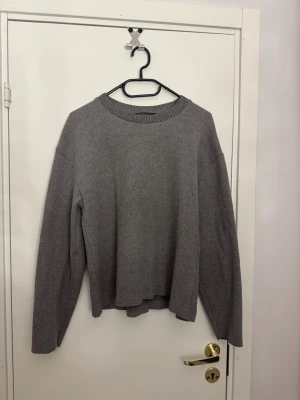 Grå fin stickad tröja - Mysig grå stickad tröja med avslappnad passform och rund halsringning. Tröjan har långa ärmar och ribbstickade detaljer vid halsen. Perfekt för lager på lager och enkel att matcha till olika outfits. Finstickat materia