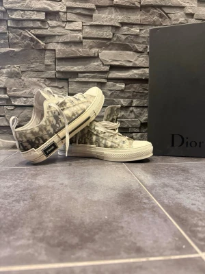 Dior B23 låga sneakers monogram - Säljer ett par Dior B23 låga sneakers med ikoniskt grått och vitt monogrammönster. Skorna har genomskinliga paneler, vita snören och en robust vit sula med svart detalj på hälen där Dior-loggan syns. Perfekta för dig som gillar exklusiv streetwear.