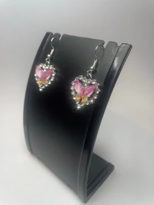 Hjärformade örhängen med fjäril - Snygga örhängen i hjärtform med en rosa och lila fjäril i mitten. Runt hjärtat sitter små klara pärlor som ger extra bling. Krokar i silverfärg. Perfekta för dig som gillar gulliga och unika accessoarer.