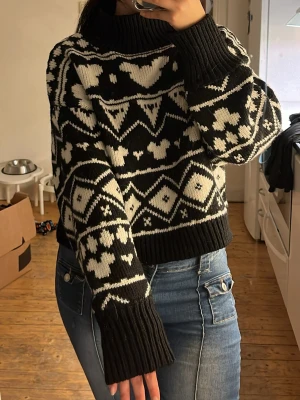 Svartvit stickad tröja med mönster - Stickad tröja i svart och vitt med grafiskt mönster och hjärtan. Tröjan har hög krage, långa ärmar och ribbade muddar vid ärmslut och nederkant. Perfekt för kalla dagar och ger en mysig, trendig look.
