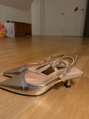 Guldiga slingback pumps med klack - Snygga pumps i metallic guld med spetsig tå och smal rem runt hälen. Skorna har en låg, smal klack och justerbart spänne. Materialet ser ut som konstläder med glansig finish. Perfekta för dig som vill sticka ut med elegans. Säljer då de ej kommer till användning, använda fåtal gånger.