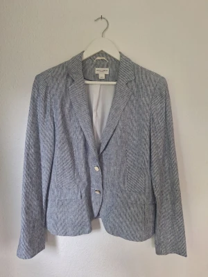 Randig kavaj från Holy and Whyte / Lindex - Snygg linnekavaj från Lindex i tunn, blåvitt smalrandig tyg med vitt foder. Klassisk modell med två knappar framtill, fickor och slits bak. Perfekt till sommaren med sitt lätta material och tidlösa design. Fodrad och med knappar även vid ärmslut.