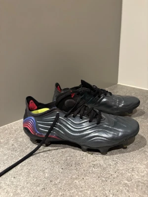 Adidas Copa Sense+ FG fotbollsskor - Snygga Adidas Copa Sense+ FG fotbollsskor i svart med vågiga detaljer och färgskiftningar i blått, rött och lila på hälen. Skorna har svarta snören, låg profil och är tillverkade i syntetmaterial för bästa bollkontroll. Perfekta för spel på naturgräs.