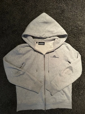  J.Lindeberg Hoodie grå - Storlek M passar S också .Snygg grå hoodie från J.Lindeberg med dragkedja framtill och justerbar huva med snören. Tillverkad i mjuk bomullsblandning och har diskret logga broderad på bröstet. Perfekt för en chill och stilren look.