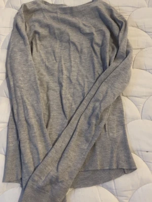 Ljusgrå stickad tröja med rund hals - En stilren ljusgrå stickad tröja med rund halsringning och långa ärmar. Tröjan har breda ribbstickade muddar vid ärmslut och nederkant, vilket ger en snygg och enkel look. Perfekt att matcha med jeans eller kjol.