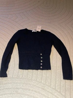 Mörkblå stickad topp med knappar - Mörkblå stickad topp i alpackablandning från Alcaa Blend. Modellen är figurnära med långa ärmar och har fem dekorativa knappar i olika färger på sidan. Perfekt för dig som gillar en enkel men ändå unik look.