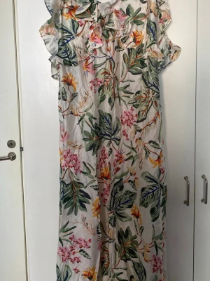 Blommig långklänning med volang - Långklänning med färgglatt blommönster i grönt, rosa, gult och orange på vit botten. Klänningen har volangdetaljer vid axlar och halsringning. Perfekt för sommarens alla tillfällen.