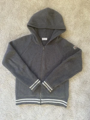 Grå Moncler hoodie med dragkedja - Snygg grå hoodie från Moncler med dragkedja framtill och huva. Tröjan har långa ärmar, ribbade muddar och vita ränder vid ärmslut och nederkant. Moncler-logga på ena ärmen ger en exklusiv touch. Perfekt för en chill och stilren look.