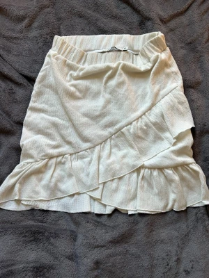 Vit kort kjol - Supersöt vit kortkjol med omlottdesign och volangdetaljer. Kjolen har en mjuk och lätt struktur, perfekt för varma dagar. Elastisk midja för skön passform och flowy känsla. Enkel att matcha till olika toppar och sneakers.