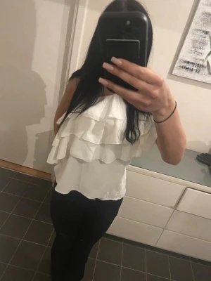 Vit oneshoulder topp med volanger - Säljer en vit oneshoulder topp med flera lager volanger framtill. Toppen är ärmlös och har en luftig passform