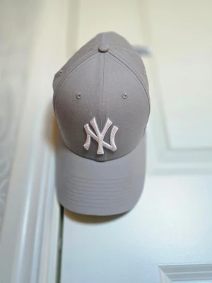 Grå New York Yankees keps - Snygg grå keps från New Era med klassisk New York Yankees-logga broderad i vitt framtill. Kepsen har böjd skärm och justerbar baksida. Tillverkad i slitstark bomull som passar perfekt till streetwear och casual outfits.