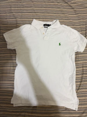 Polo ralph lauren polo pike tröja - Klassisk vit pikétröja från Polo Ralph Lauren med grönt broderat logotyp på bröstet. Tröjan har korta ärmar, krage och knappar framtill. Tillverkad i bomull och har en clean, tidlös look som alltid funkar. Sitter perfekt som S