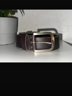 Mörkbrunt skärp från Tom Tailor - Snyggt mörkbrunt bälte från Tom Tailor i äkta skinn. Klassisk design med silverfärgat metallspänne och robust känsla. Perfekt accessoar för att lyfta din outfit med en tidlös touch.