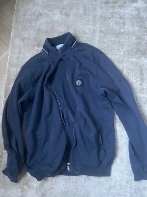 Stone Island full zip - En marinblå Stone Island fullzip i storlek M/ L. Är i ganska bra skick med 2st små fläckar på ena armen som inte syns vid användning. Skriv vid minsta lilla fundering!