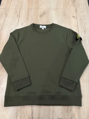 Olivgrön sweatshirt från Stone Island - Säljer en olivgrön sweatshirt från Stone Island med klassisk rund hals och ribbade muddar. Tröjan har den ikoniska Stone Island-loggan på vänster ärm. Perfekt för dig som gillar streetwear och enkel stil.