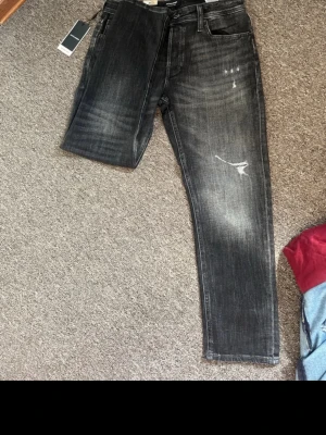 Svarta slitna jeans med tvättad look - Säljer ett par svarta jeans med tvättad och sliten look, raka ben och detaljerade slitningar på framsidan. Jeansen har klassisk femficksmodell och knappgylf. Perfekta för dig som gillar en avslappnad och trendig stil.