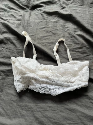 Vit spetsbralette med smala band - Säljer en vit bralette i mjuk spets med smala, justerbara axelband. Braletten har ett fint spetsmönster och en dekorativ kant nedtill. Perfekt för dig som gillar en enkel och bekväm stil. Den är i S men passar xs och neråt också.