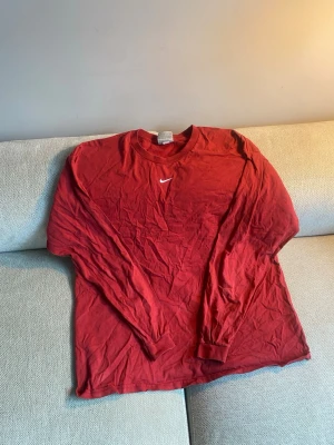 Röd långärmad tröja från Nike - Snygg röd långärmad tröja från Nike med klassisk vit swoosh-logga på bröstet. Tröjan har rund halsringning och ribbade muddar vid ärmsluten. Perfekt för en avslappnad och sportig stil.
