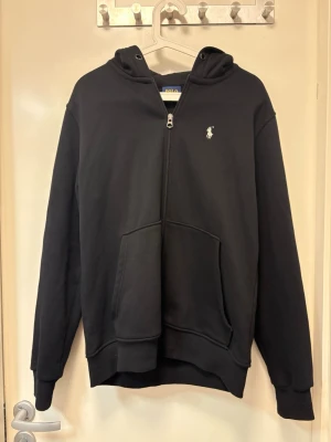 Svart hoodie från Polo Ralph Lauren - Klassisk svart hoodie från Polo Ralph Lauren med dragkedja framtill och vit broderad logga på bröstet. Hoodien har stor känguruficka och huva. Tillverkad i mjukt bomullsmaterial som känns skönt mot huden. Perfekt för en avslappnad och stilren look.