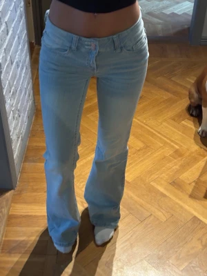 Ljusblå bootcut jeans med rosa söm - Säljer ett par ljusblå bootcut jeans med låg midja och rosa dekorationssöm på bakfickan. Jeansen har klassisk femficksdesign och är tillverkade i mjukt denimtyg. Perfekta för en avslappnad och trendig look.