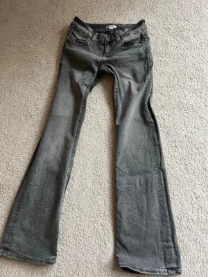 Grå bootcut jeans med detaljer - Säljer ett par grå bootcut jeans med snygga slitningar och kontrastsömmar. Jeansen har fem fickor och cool knappdetalj fram. Perfekta för dig som gillar en avslappnad men trendig look.