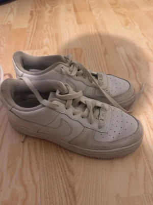 Nike Air Force 1 vita sneakers - Klassiska Nike Air Force 1 sneakers i helvitt läder med perforerad tåbox och vit sula. Skorna har lågt skaft, snörning och den ikoniska swoosh-loggan på sidorna. Perfekta för en clean och stilren look.