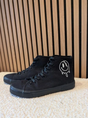 Svarta high top sneakers med smiley - Säljer ett par svarta high top sneakers i canvas med svart gummisula och snörning. På utsidan finns en vit smiley med drip-effekt som ger en cool streetkänsla. Perfekta för dig som gillar unik och edgy stil.