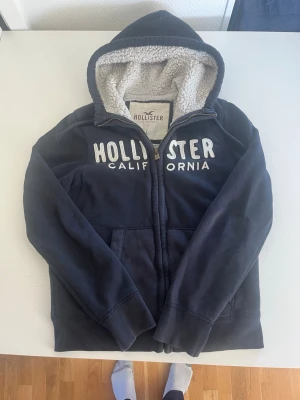 Hollister California zip hoodie  - Snygg marinblå hoodie från Hollister med dragkedja och stor vit broderad logga på bröstet. Insidan av huvan är fodrad med mjuk, vit teddy för extra komfort. Klassisk passform och två fickor framtill. Perfekt för en avslappnad och cool stil.