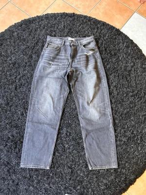 Grå mom jeans med slitningar - Säljer ett par grå mom jeans med hög midja och raka ben. Jeansen har slitningar vid fickan och vid bensluten för en trendig look. Klassisk femficksmodell i kraftig denim, perfekta till vardagsstilen.