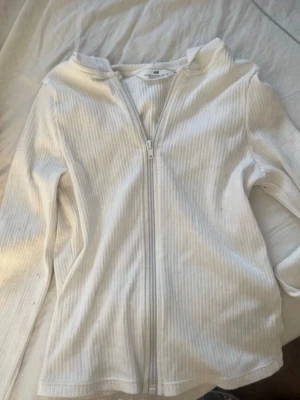 Vit ribbad ziptop från H&M - Vit ribbad långärmad topp från H&M med dragkedja framtill och huva. Perfekt för en chill och stilren look men också för träning 🎾. Materialet är mjukt och stretchigt för extra komfort. STORLEK 158/164 men funkar som XXS om man är lite kortare❤️💐