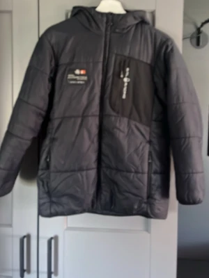 Svart dunjacka från Sail Racing - Säljer en svart dunjacka från Sail Racing med huva och dragkedja framtill. Jackan har en bröstficka med logga och patch med texten 'Sail Antarctica'. Perfekt för kalla vinterdagar och har en sportig look med diskreta detaljer. Bara att höra av sig vid funderingar!