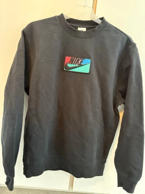 Svart Nike sweatshirt med färgglad logga - Svart sweatshirt från Nike med en färgglad broderad logga på bröstet i rött, blått och turkost. Tröjan har rund halsringning, långa ärmar och ribbade muddar vid ärmslut och nederkant. Perfekt för en sportig och avslappnad stil.