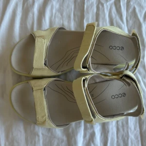 Beige gul sandaler från ECCO - Snygga och bekväma sandaler från ECCO i beige läder med justerbara kardborreband. Sulan är vit med grå detaljer och ni har bra grepp. Perfekta för sommaren och långa promenader. Klassisk design med öppen tå och häl.