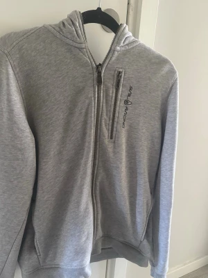 Grå hoodie från Sail Racing - Snygg grå hoodie från Sail Racing med dragkedja framtill och en extra bröstficka med zip. Hoodien har broderad logga på bröstet och en detalj på huvan. Perfekt för en avslappnad och sportig stil.
