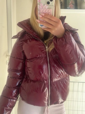 Vinröd glansig pufferjacka - Säljer en vinröd pufferjacka med glansig finish och hög krage. Jackan har dragkedja framtill och breda kanaler som ger en riktigt puffig look. snygg till streetwear-stilen.