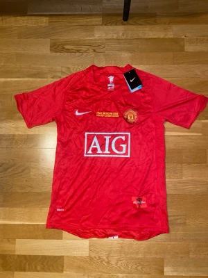 Manchester United 2008 UCL Final - Röd Manchester United matchtröja från Nike med AIG-logga på bröstet och klubbmärke. Tröjan har korta ärmar, rund hals och detaljer om Champions League-finalen i Moskva 2008. Tillverkad i lätt och ventilerande material, perfekt för fotboll eller träning.