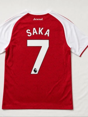 Arsenal matchtröja Saka 7 Adidas - Röd och vit Arsenal matchtröja från Adidas med Saka och nummer 7 på ryggen. Tröjan har klubbmärke och Adidas-logga på bröstet, samt sponsortryck. Tillverkad i lätt och ventilerande material, perfekt för fotboll eller träning.
