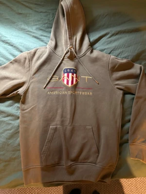 Beige hoodie från GANT med brodyr - Snygg beige hoodie från GANT med broderad logga och texten 'American Sportswear' på bröstet. Klassisk känguruficka framtill och justerbar huva med dragsko. Perfekt för en avslappnad och sportig stil.