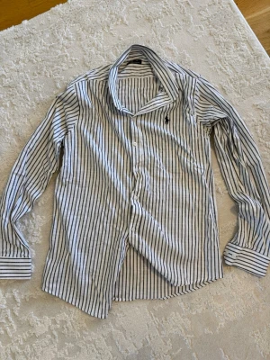 Randig skjorta från Ralph Lauren - Klassisk vit skjorta med svarta ränder från Ralph Lauren. Skjortan har lång ärm, knappar framtill och broderad logga på bröstet. Perfekt för en stilren look och tillverkad i mjuk bomull.