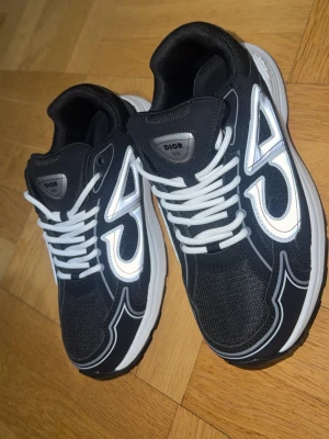 Svarta Dior sneakers med vita detaljer - Snygga svarta Dior sneakers med vita och reflekterande detaljer, vita skosnören och tjock vit sula. Skorna har en sportig design med mesh och syntetmaterial samt tydlig Dior-logga på plösen och insidan. Perfekta för dig som gillar exklusiva och trendiga sneakers.