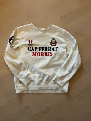 Vit sweatshirt från Morris Cap Ferrat (beskrivning) - Vit sweatshirt från Morris med tryck 'Cap Ferrat' i svart och 'Morris' i rött på bröstet. Broderad patch och emblem framtill samt tryckt siffra på ärmen. Rund halsringning och ribbade muddar. Perfekt för en sportig och stilren look. Säljer gärna via instabox!!