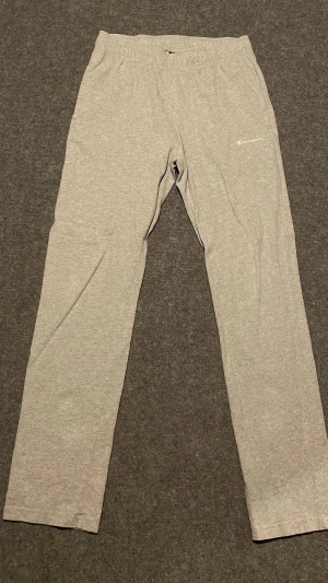 Champion Grå mjukisbyxor - Gråa mjukisbyxor från Champion med elastisk midja och raka ben. Byxorna har en diskret logga på vänster lår och är tillverkade i mjukt bomullsmaterial. Perfekta för chill eller träning.