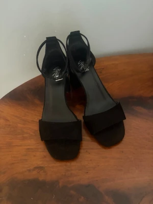 Svarta pumps med blockklack - Stilrena svarta pumps från Shoe All med öppen tå, bred rem över foten och justerbar ankelrem. Skorna har en bekväm blockklack och är tillverkade i mockaliknande material. Perfekta för dig som vill ha en klassisk och trendig look.