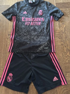 Real Madrid träningsset Adidas stl: 11-12 152 cm - Snyggt svart träningsset från Adidas och Real Madrid med rosa detaljer och tryck. Tröjan har mönstrad yta, rosa ränder på sidorna och klubbmärke på bröstet. Shortsen har matchande rosa ränder och loggor. Perfekt för fotbollsträning eller sport.