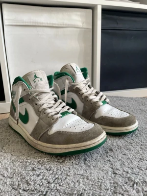 Air Jordan 1 Mid vit/grön/grå - Snygga Air Jordan 1 Mid sneakers i vitt läder med gråa mockadetaljer och gröna accenter på swoosh, häl och logga. Klassisk siluett med perforerad tå och gröna detaljer på sulan. Perfekta för dig som gillar streetwear och vill sticka ut. Ny pris 3000 kr. Det har blivit två knutar på ena skosnöret tyvärr men funkar ändå