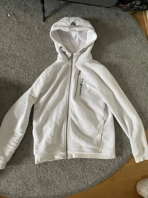 Vit hoodie från sail racing - Snygg vit hoodie från sail racing med dragkedja framtill och en bröstficka med logga. Hoodien har en stor huva med snörning och är tillverkad i mjukt material, perfekt för en avslappnad stil.