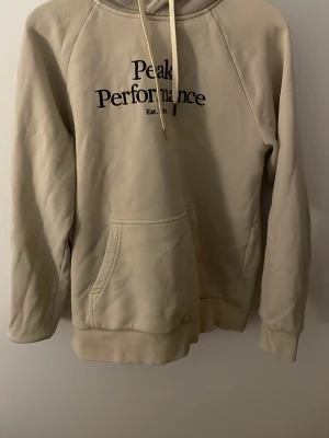 Beige hoodie från Peak Performance - Snygg beige hoodie från Peak Performance med svart logga och text på bröstet. Klassisk känguruficka framtill, ribbade muddar och dragsko i huvan. Perfekt för en chill och avslappnad stil. Mjuk bomullskänsla och riktigt skön passform.