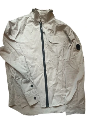 Beige overshirt från C.P. Company - Stilren beige/grå overshirt från C.P. Company med svart dragkedja, bröstficka med lock och ikonisk linsdetalj på ärmen. Overshirten har klassisk krage och är tillverkad i ett lätt material som passar perfekt för lager-på-lager-stil. Storlek L men passar mer M. 